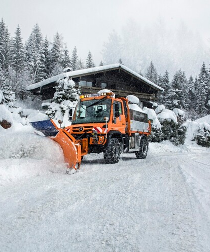 Ein orangefarbener Unimog räumt Schnee vor einem Haus.