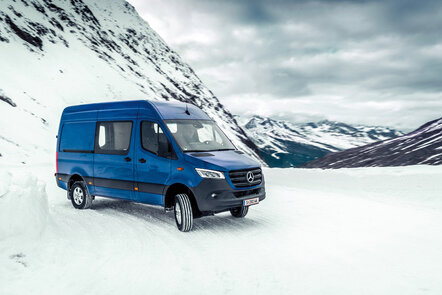 Ein blauer Mercedes-Benz Transporter fährt durch schneebedeckte Berglandschaft.