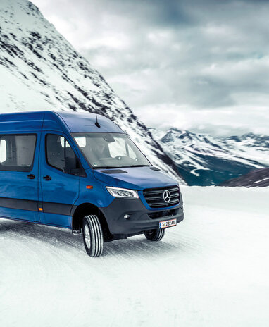 Ein blauer Mercedes-Benz Transporter fährt durch schneebedeckte Berglandschaft.