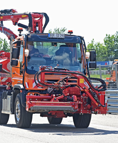Ein oranger Unimog mit einem roten Mäharm fährt auf einer Straße.