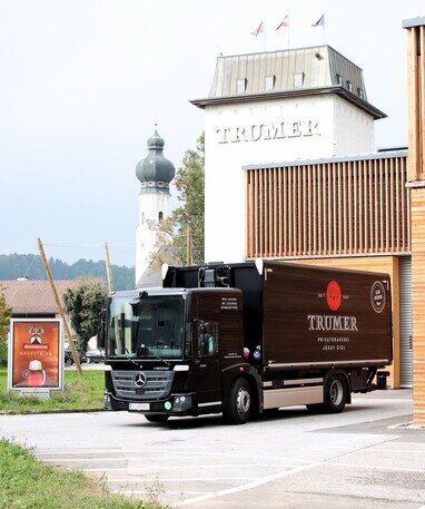 Der Mercedes-Benz eEconic mit dem Logo der Trumer Brauerei vor dem Gebäude der Brauerei.