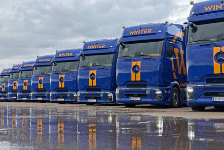 Acht blaue Mercedes-Benz Actros Lkw nebeneinander, die sich am nassen Boden spiegeln.