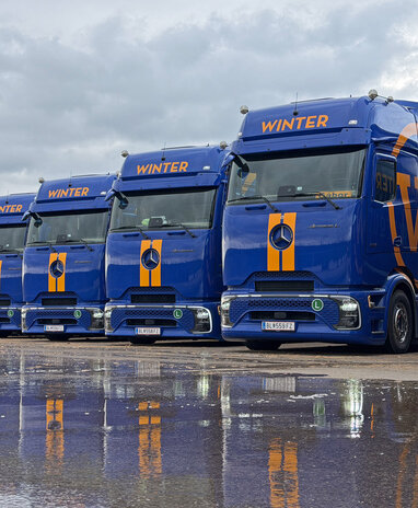 Acht blaue Mercedes-Benz Actros Lkw nebeneinander, die sich am nassen Boden spiegeln.