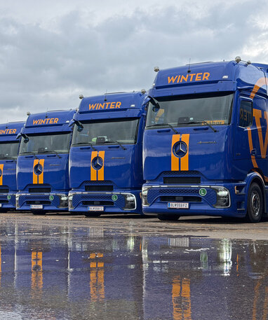 Acht blaue Mercedes-Benz Actros Lkw nebeneinander, die sich am nassen Boden spiegeln.