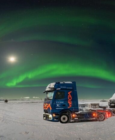 Blauer eActros600 mit orangem VEGA-Logo mit Sattelzug auf einer schneebedeckten Fläche unter grünem Polarlicht.