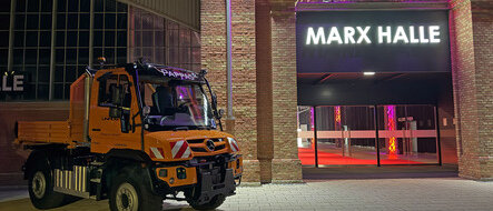 Ein orangefarbener Mercedes-Benz Unimog steht vor der beleuchteten Marx Halle in einer nächtlichen Szene.