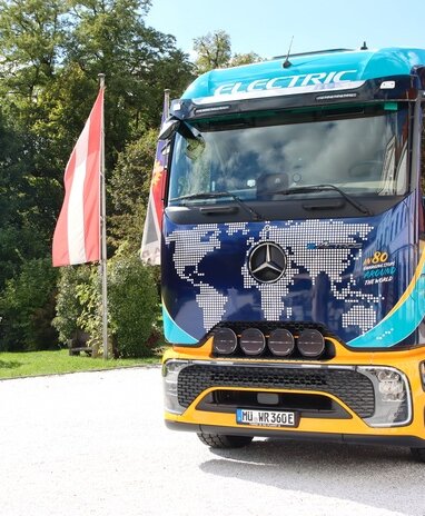 Ein elektrischer Mercedes-Benz Lkw mit Weltkarten-Design und der Aufschrift 'ELECTRIC' steht neben einer Fahne.
