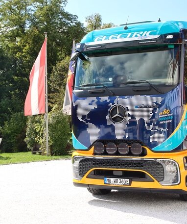 Ein elektrischer Mercedes-Benz Lkw mit Weltkarten-Design und der Aufschrift 'ELECTRIC' steht neben einer Fahne.
