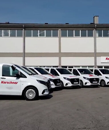 6 weiße Vito Tourer mit Kerschner-Branding stehen nebeneinander vor dem Kerschner Firmengebäude.
