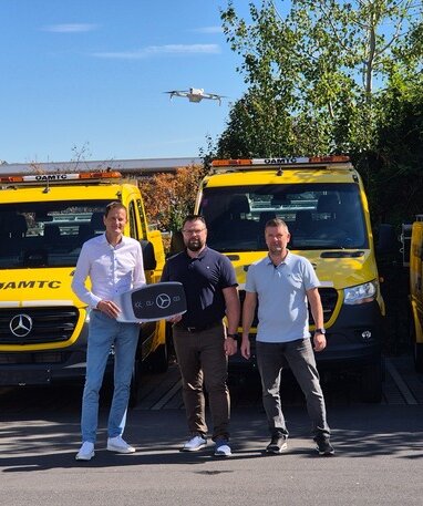 Drei Männer vor ÖAMTC-Pannenfahrzeugen mit Mercedes-Benz-Logo bei der Schlüsselübergabe.