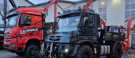 Ein roter Lkw und ein schwarzer Unimog mit Kranaufbauten auf einem Betriebshof.