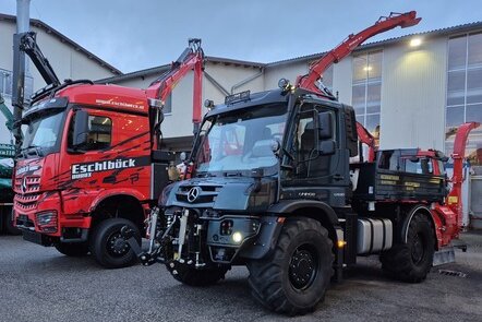 Ein roter Lkw und ein schwarzer Unimog mit Kranaufbauten auf einem Betriebshof.