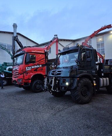 Ein roter Lkw und ein schwarzer Unimog mit Kranaufbauten stehen in der Abenddämmerung nebeneinander.