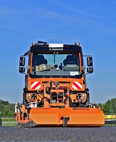 Eine orangefarbene Unimog mit Kehraufsatz von Kahlbacher auf einer Straße.