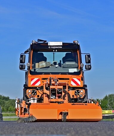 Eine orangefarbene Unimog mit Kehraufsatz von Kahlbacher auf einer Straße.
