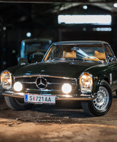 Ein dunkler, klassischer Mercedes-Benz 250 SL mit eingeschalteten Scheinwerfern in einer Werkstatt.