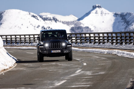 Ein schwarzer Jeep Wrangler fährt auf einer Bergstraße durch eine schneebedeckte Landschaft.