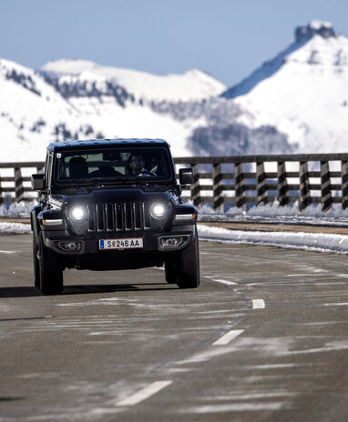 Ein schwarzer Jeep Wrangler fährt auf einer Bergstraße durch eine schneebedeckte Landschaft.