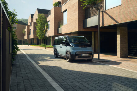 Blauer Kia PV5 Passenger in schräger Frontansicht zwischen modernen Wohnhäusern.
