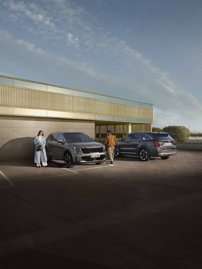 Zwei Kia Sorento in schräger Ansicht stehen vor einer modernen Fassade mit zwei Personen neben den Fahrzeugen.