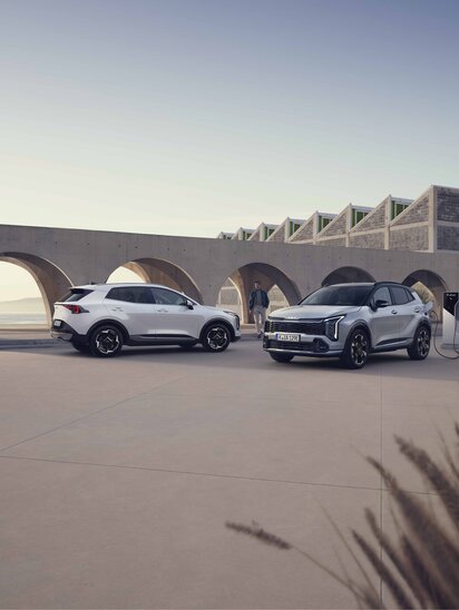 Zwei Kia Sportage stehen auf offenem Platz vor moderner Architektur, einer in schräger Front-, der andere in schräger Heckansicht.