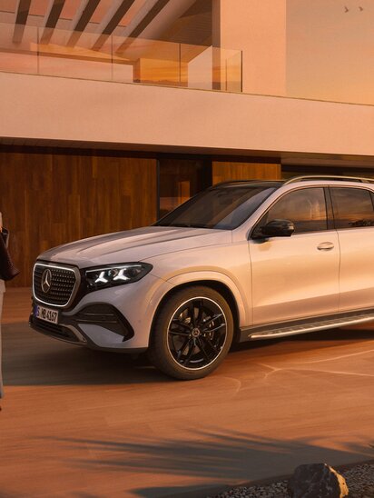 Elegante Frau steht neben einem geparkten Mercedes-Benz GLE vor einem modernen Haus bei Sonnenuntergang. | © Mercedes-Benz AG