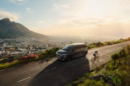 Der Mercedes-Benz VLE fährt auf einer Bergstraße neben einem Radfahrer; im Hintergrund ist eine Stadtlandschaft bei Sonnenaufgang zu sehen. | © Mercedes-Benz AG
