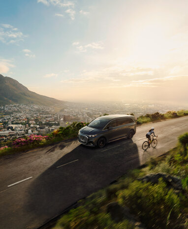 Der Mercedes-Benz VLE fährt auf einer Bergstraße neben einem Radfahrer; im Hintergrund ist eine Stadtlandschaft bei Sonnenaufgang zu sehen. | © Mercedes-Benz AG