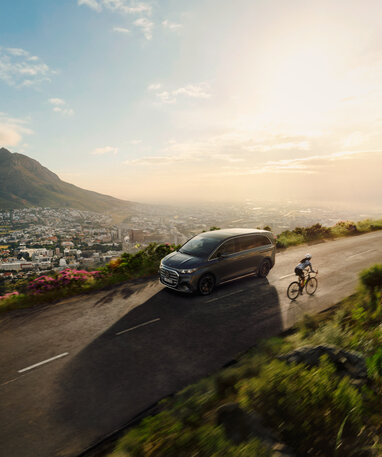 Der Mercedes-Benz VLE fährt auf einer Bergstraße neben einem Radfahrer; im Hintergrund ist eine Stadtlandschaft bei Sonnenaufgang zu sehen. | © Mercedes-Benz AG