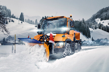 Ein Mercedes-Benz Unimog U 323 Schneepflug räumt Schnee von einer Straße in einer winterlichen Berglandschaft.