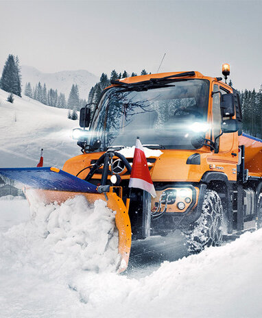 Ein Mercedes-Benz Unimog U 323 Schneepflug räumt Schnee von einer Straße in einer winterlichen Berglandschaft.