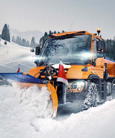Ein Mercedes-Benz Unimog U 323 Schneepflug räumt Schnee von einer Straße in einer winterlichen Berglandschaft.